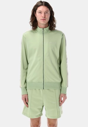 Tuta leggero verde chiaro con giacca con zip e pantaloni corti. Presenta un colletto alto, strisce laterali e una trama liscia. Branding minimale visibile.