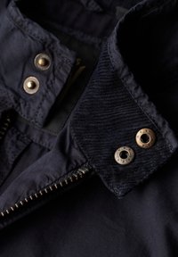 Granatowa kurtka z teksturowanym kołnierzem, metalowymi zatrzaskami i suwakiem, wykonana z trwałej tkaniny o gładkim wykończeniu.