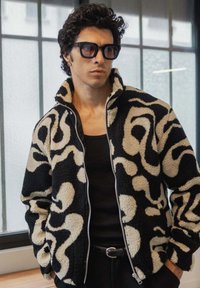 Veste en polaire noire et crème texturée avec un motif abstrait en swirl, fermeture éclair intégrale et col montant. Portée sur un débardeur noir.