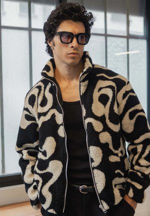 Veste en polaire noire et crème texturée avec un motif abstrait en swirl, fermeture éclair intégrale et col montant. Portée sur un débardeur noir.