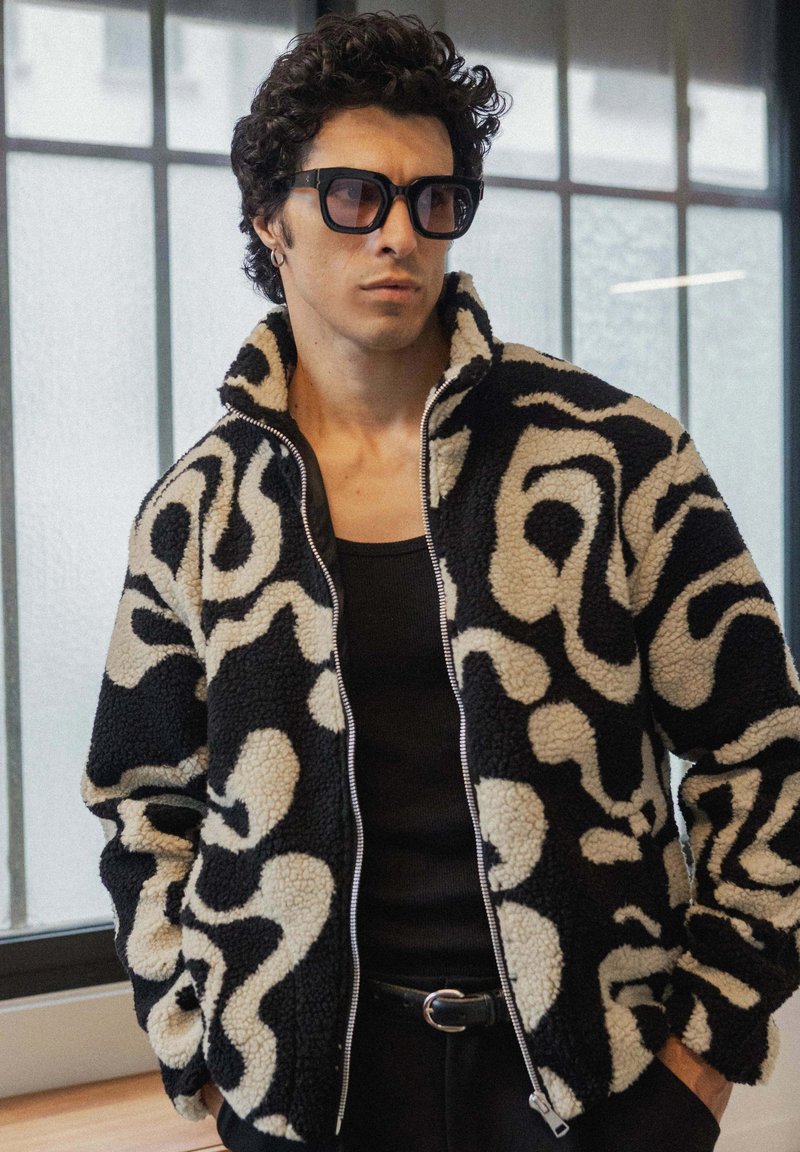 Veste en polaire noire et crème texturée avec un motif abstrait en swirl, fermeture éclair intégrale et col montant. Portée sur un débardeur noir.