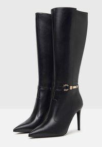 Bottes en cuir noires montant jusqu'au genou, avec des bouts pointus, des talons hauts fins et des sangles décoratives avec boucle dorée autour de la cheville.