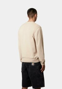 Suéter beige de punto con cuello redondo, puños y dobladillo acanalados, combinado con pantalones cortos oscuros que tienen bolsillos traseros y un parche con el logo.