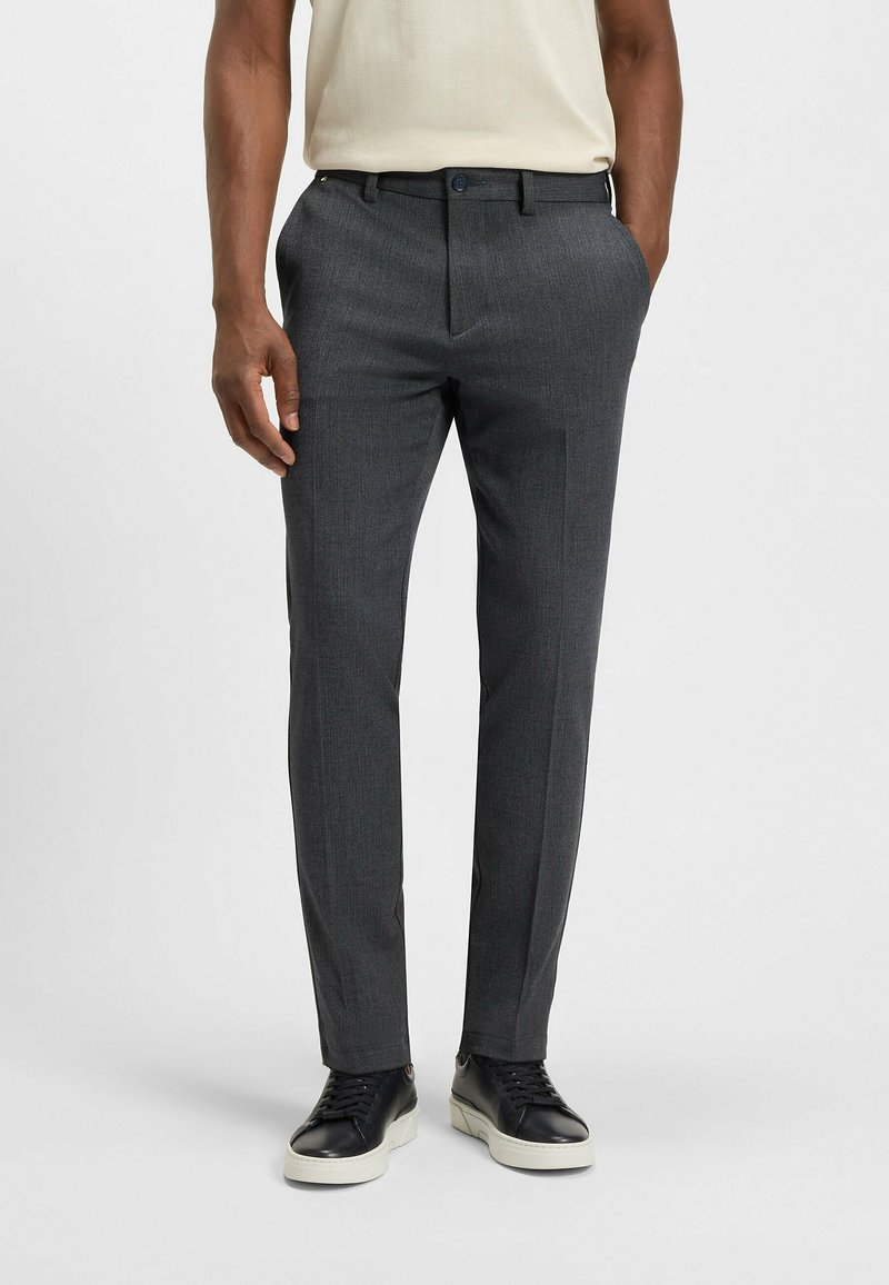 Homme portant un pantalon habillé slim gris foncé, des baskets en cuir noir avec des semelles blanches, et une chemise claire rentrée à l'intérieur.