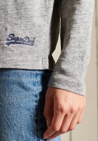 Harmaa pitkähihaan paita, jossa on pyöreä pääntie. Paidassa on sininen "SuperDry"-logo helman kohdalla. Yhdistetty sinisiin farkkuihin, jotka näyttävät teksturoidulta kankaalta.
