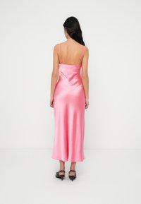 Ροζ σατέν μίντι φόρεμα με χαμηλή πλάτη,  λεπτές ρυθμιζόμενες τιράντες και λεία υφή, που ρέει μέχρι τους αστραγάλους. Φορεμένο με μαύρες γόβες.