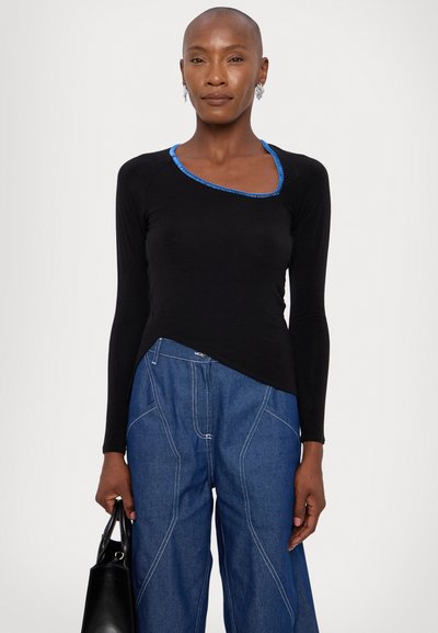 Top lungo nero a maniche lunghe con accento blu sul collo, caratterizzato da un orlo asimmetrico unico. Abbinato a pantaloni in denim blu a gamba larga.
