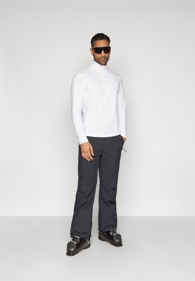 Λευκή ζακέτα με μακρύ μανίκι και φερμουάρ, μαύρο παντελόνι σκι και μαύρες μπότες σκι. Μοντέλο που φορά σκούρα γυαλιά ηλίου, στέκεται μπροστά σε γκρι φόντο.