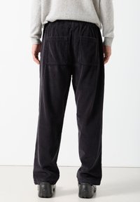 Osoba koja nosi široke crne manšetrirane hlače s velikim stražnjim džepovima i crnim cipelama, stojeći na bijeloj pozadini.