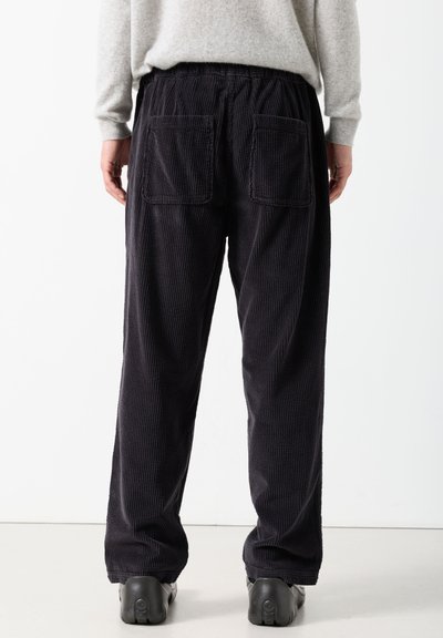 Personne portant un pantalon en velours côtelé noir ample avec de grandes poches arrière et des chaussures noires, debout devant un fond blanc.
