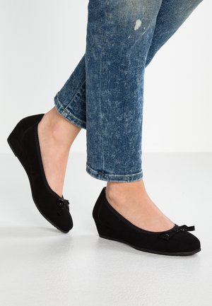 Fødder iført sorte kilehæl-ballerinasko med små sløjfer, kombineret med blå, op rullede denim jeans på en lys gulvbaggrund.