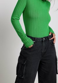 Haut vert à manches longues côtelées associé à un pantalon cargo noir avec poches latérales et coupe décontractée. Tricot texturé et matériaux en denim apparents.