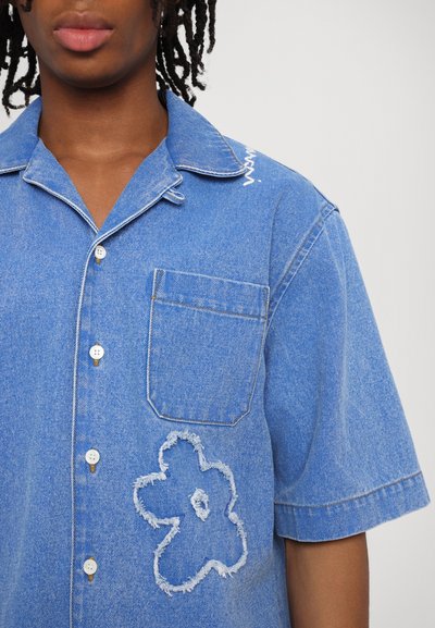 Zila denim krekls ar īsām piedurknēm, norādītu apkakli, krūšu kabatu un nodilušu ziedu dizainu priekšpusē. Balti šuvumi uzsver.