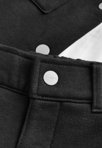 Čierna látka s hladkou textúrou, zdobená strieborným cvokem s označením "ALLSAINTS" a kontrastným bielym panelom v abstraktnom dizajne.