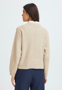 Béžový pletený sveter s rebrovanou textúrou a spustenými ramenami, s krátkym lemom a mäkkým, vrstveným bielym golierom pod ním.