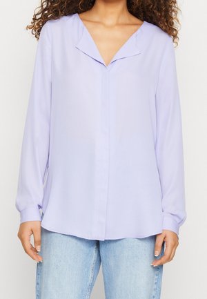 Kvinde iført en lyse lilla langærmet bluse med en V-formet krave og lyseblå jeans, stående mod en ensfarvet baggrund.