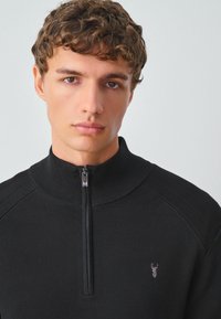 Maglione nero a collo alto con zip a un quarto, texture a costine e un piccolo disegno ricamato sul petto sinistro. Vestibilità casual.