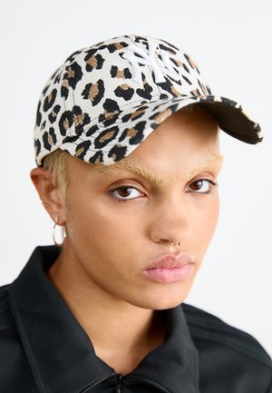 Personne à la peau brun clair portant une veste noire, une casquette de baseball à imprimé léopard avec logo blanc "NY", une boucle d’oreille en anneau et un piercing au septum.