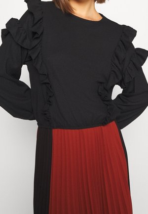 Kobieta ubrana w czarną, teksturowaną bluzkę z falbanianymi rękawami oraz plisowaną spódnicę z czerwonymi i czarnymi pionowymi panelami.