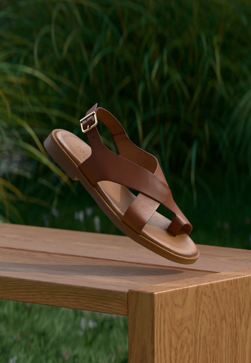 Καφέ δερμάτινο σανδάλι με λουρί και πόρπη αιωρείται πάνω από ξύλινο τραπέζι, πράσινα φυτά είναι θολά στο φόντο.