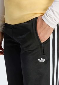 Black athletic pants with white three-stripe design on sides and zippered pocket. Features a small white logo on the thigh area.