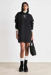 Svart sweatshirtklänning med en dekorativ "CK"-logotyp, ribbade ärmslut och en lös passform. Kombinerad med knähöga strumpor och kraftiga svarta stövlar.