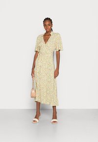 Vestido envelopado floral em amarelo claro com padrões verdes e rosas, mangas curtas e uma fenda lateral. Usado com sandálias de salto bege e uma bolsa pequena.