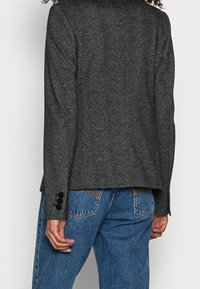 Grauer Fischgrät-Blazer mit Kragen, langen Ärmeln und drei Knöpfen an jedem Ärmelabschluss, kombiniert mit blauen Jeans.
