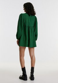 Groene geruite mini-jurk met poffende mouwen, verzamelde taille en een uitlopende rok. Draagt met zwarte enkellaarsjes. Effen witte achtergrond.