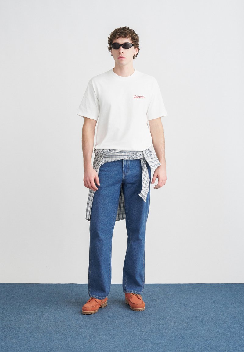 White t-shirt with red logo, plaid shirt tied at the waist, blue jeans, and orange shoes. Model wearing black sunglasses, standing on a blue floor.