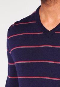 Pull en maille bleu marine à col en V avec des rayures horizontales rouges et grises. Texture lisse avec une coupe ajustée.