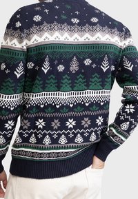 Marineblauer Pullover mit winterlichen Mustern in Grün, Weiß und Grau, darunter Schneeflocken, Kiefern und geometrische Formen, getragen mit weißen Hosen.