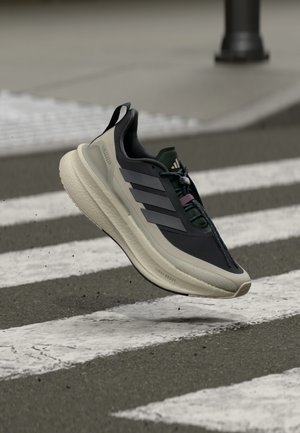 Svart løpesko med grå detaljer, med tre striper, teksturert såle og en polstret mellomsåle, plassert over et fotgjengerfelt.