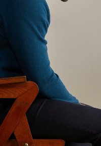 Personne en pull bleu et pantalon noir assise de côté sur une chaise en bois, devant un fond beige uni.