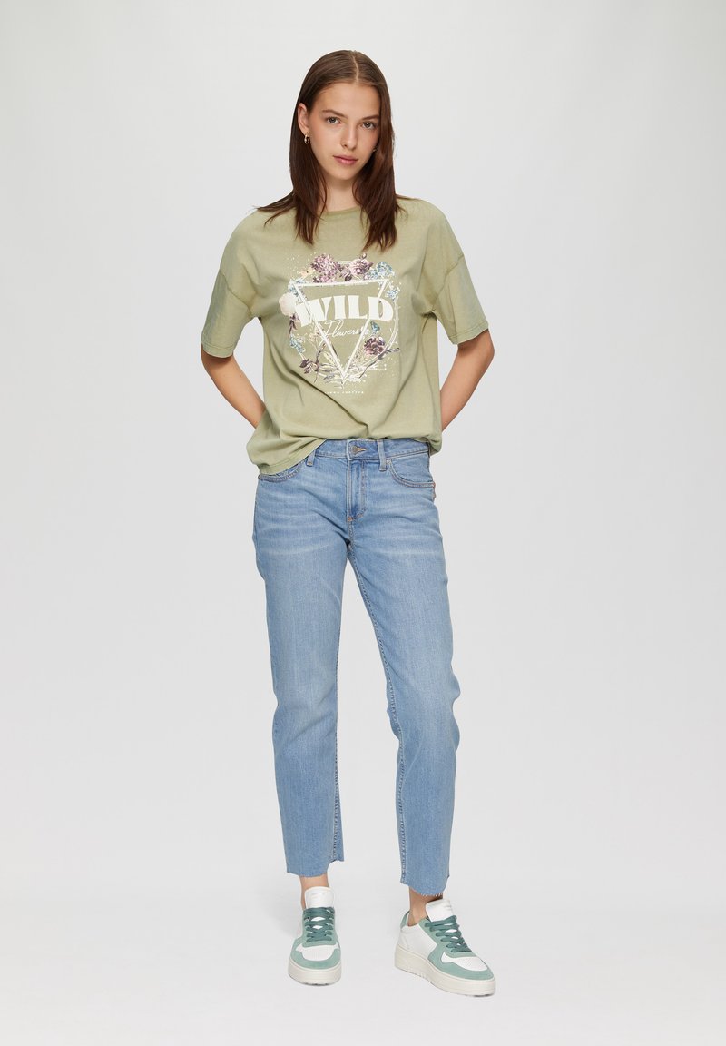 Olijfgroene oversized t-shirt met bloemenprint, gecombineerd met lichtblauwe straight-leg jeans en witte sneakers met groene accenten.