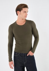 Olivenfarget langermet skjorte i en tettsittende design med rund hals, kombinert med mørke denimjeans med kontrastsømmer.