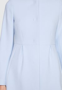 Primo piano di un vestito azzurro chiaro a maniche lunghe con cuciture su misura e vita aderente, caratterizzato da leggere pieghe sui fianchi.