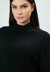 Schwarzer Strickpullover mit hohem, geripptem Kragen und strukturierten Zopfmustern. Der Stoff wirkt weich und hat ein strukturiertes Design.