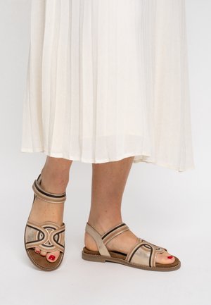 Beige Sandalen mit mehreren Riemen, runden Designelementen und brauner Sohle. Weiche Wildledertextur. Getragen zu einem plissierten, hellfarbenen Kleid.