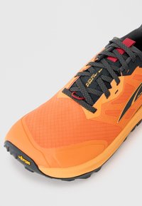 Oranssi ja musta ulkoilukenkä mustilla nauhoilla, punaisella nauhakoukulla ja Vibram-pohjan logolla valkoista taustaa vasten.