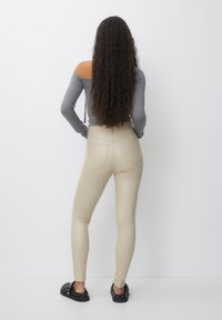 Leggings extensibles beiges à taille haute, avec deux poches arrière et une texture lisse et brillante, associés à un haut à manches longues gris.