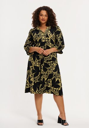 Kvinde iført sort og gul blomsterprint midi-kjole med trekvartlange ærmer og sorte blokhældte sko med åben tå, stående med hænderne foldet.