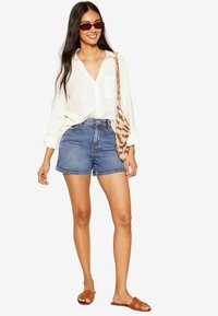 Shorts in denim blu medio con vita alta, abbinati a una camicia bianca con bottoni e sandali marroni, accessoriati con una borsa con motivo.