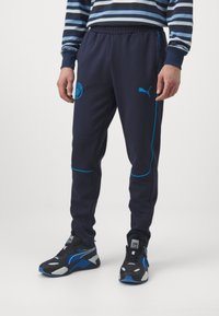 Jūras spēku jogger bikses ar gaiši ziliem akcentiem un Puma logo. Iespējas ietver elastīgu jostasvietu un ribotiem apročām. Materiāls ir mīksts, sportisks audums.