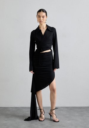 Top negru cu mâneci lungi, asociat cu o fustă neagră plisată cu un tiv asimetric. O pereche de sandale cu barete negre completează ținuta.