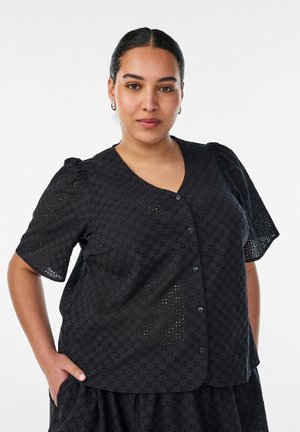 Kvinde med mørkt hår samlet bagud iført sort øjenhuls-knap-bluse og matchende nederdel, poserer med den ene hånd i lommen mod hvid baggrund.