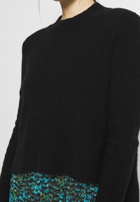 Kvinna klädd i en svart, fluffig långärmad tröja som matchas med en blå och grön mönstrad kjol, visad från axlarna till höfterna.