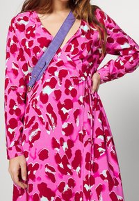 Pink wrap dress featuring a bold red and teal pattern, long sleeves, and a tie detail at the waist. Accented with a purple strap.