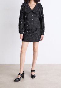 Completo nero con texture, composto da una camicia con colletto e bottoni arricchita da dettagli in perle e una gonna abbinata, abbinato a scarpe con tacco nere lucide.