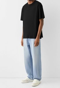 Henkilö, jolla on yllään löysä musta t-paita, vaaleansiniset leveälahkeiset farkut ja valkoiset tennarit, seisoo yksinkertaista valkoista taustaa vasten.